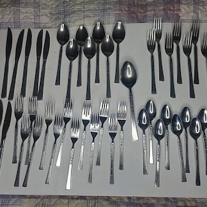 May include: A large collection of stainless steel flatware arranged on a white surface, including various dinner knives, dinner forks, salad forks, and spoons. The set features a mix of plain handles and pieces with decorative embossed patterns.