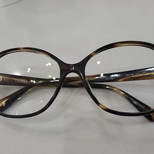 May include: Tortoiseshell-patterned eyeglasses with a rounded frame shape. The inner temple arms feature the text Oliver Peoples, 50 18 140, and Polarized. The frames have a glossy finish and clear lenses, showcasing a classic design with visible hinges.