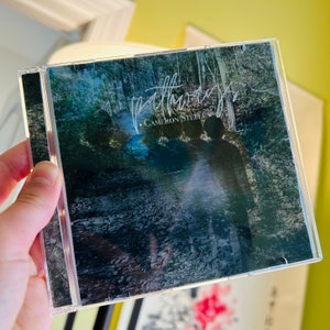 Puede incluir: Una caja de CD con fondo negro y una imagen borrosa de un camino en el bosque. El texto "Pathways" est&aacute; escrito en cursiva blanca con el nombre del artista "Cameron Steele" debajo.