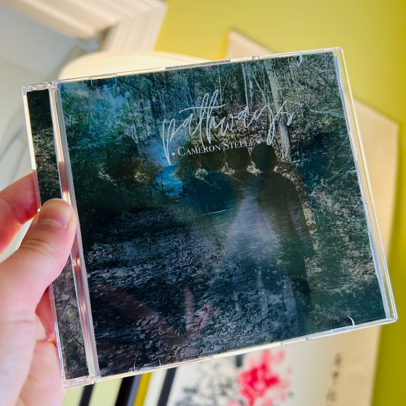 Puede incluir: Una caja de CD con fondo negro y una imagen borrosa de un camino en el bosque. El texto "Pathways" est&aacute; escrito en cursiva blanca con el nombre del artista "Cameron Steele" debajo.