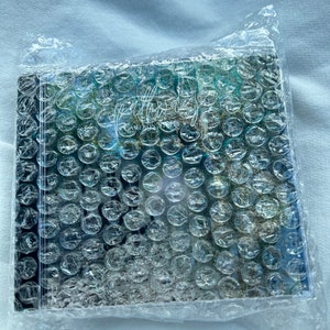 Puede incluir: Una caja de CD con una cubierta de pl&aacute;stico de burbujas. La caja de CD tiene un dise&ntilde;o abstracto azul y verde con el texto "Afterglow" y "Cameron Steele" en la parte delantera.