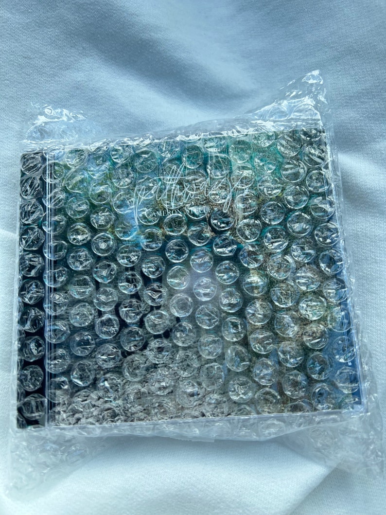 Puede incluir: Una caja de CD con una cubierta de pl&aacute;stico de burbujas. La caja de CD tiene un dise&ntilde;o abstracto azul y verde con el texto "Afterglow" y "Cameron Steele" en la parte delantera.