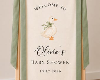 May include: A cream-colored fabric banner hanging from a light wood stand, featuring a watercolor duck illustration, sage green ribbon bows, and the text: WELCOME TO Olivia's BABY SHOWER 10.17.2026. The sign is printed with black serif and script lettering.