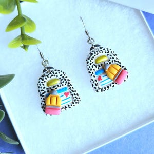 May include: A pair of dangle earrings shaped like miniature backpacks with a black and white speckled pattern. Each backpack features a small yellow pencil with a pink eraser, a gold-colored top handle, and a blue lined notebook detail with a red heart.