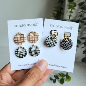 Pode incluir: Brincos redondos com padrão de grade em castanho, preto e branco. Os brincos estão em cartões brancos com o texto "STUDIONINORI" e "www.studioninori.com". Um par tem argolas douradas. Têm um buraco na parte superior.