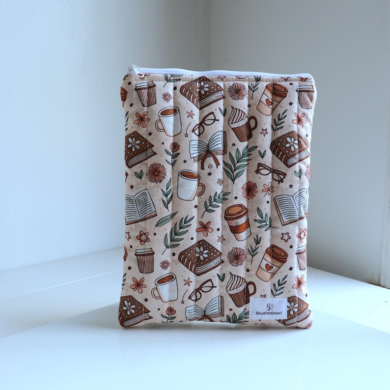 May include: A rectangular quilted fabric pouch featuring a repeating pattern of books, coffee cups, eyeglasses, and small flowers in shades of brown, cream, and green. The pouch has a white zipper closure and a small white label reading Studioninori.