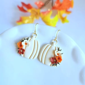 Puede incluir: Un par de pendientes colgantes con dijes en forma de calabaza blanca. Cada calabaza está decorada con pequeñas flores coloridas en tonos naranja, rojo y blanco, unidas a ganchos de metal dorado para orejas perforadas.