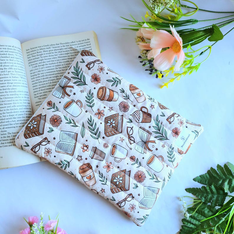 May include: A rectangular quilted fabric pouch featuring a repeating pattern of open books, coffee cups, eyeglasses, and small flowers in shades of brown, green, and orange on a cream background, with a small white tag labeled BookishBound.