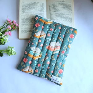 May include: A rectangular quilted fabric sleeve featuring a teal background with a repeating pattern of sleeping cats resting on stacks of books, interspersed with pink flowers and green leaves. A small white label reads Studio Inari.