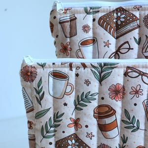 May include: Two quilted zippered pouches featuring a repeating pattern on a light beige background. The print includes brown books with white flowers, white coffee mugs, brown to-go cups, eyeglasses, green leaves, and small orange flowers.