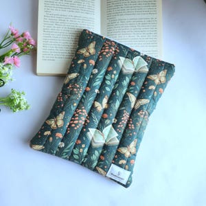 May include: A rectangular quilted fabric sleeve featuring a dark green background with a repeating pattern of butterflies, open books, and small orange and white flowers. A small white rectangular label with the text Studioinori is attached.