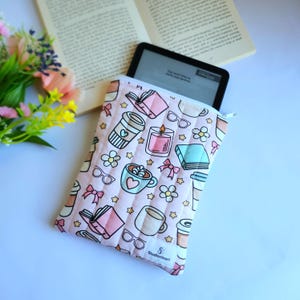May include: A quilted fabric sleeve with a light pink background featuring a repeating pattern of books, coffee cups, candles, glasses, flowers, and bows. A black e-reader is partially inserted into the sleeve, which has a white label reading StudioNiori.