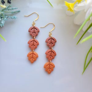 May include: A pair of dangle earrings featuring three leaf-shaped charms in shades of orange and brown. The earrings have gold-colored hooks. The leaves are arranged vertically, creating a fall-themed accessory.
