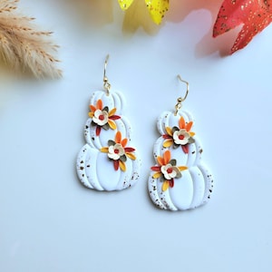 May include: A pair of dangle earrings featuring stacked white pumpkin shapes with gold-toned speckles. Each pumpkin is adorned with small, layered floral accents in shades of orange, yellow, and dark green, attached to gold-toned metal hooks.