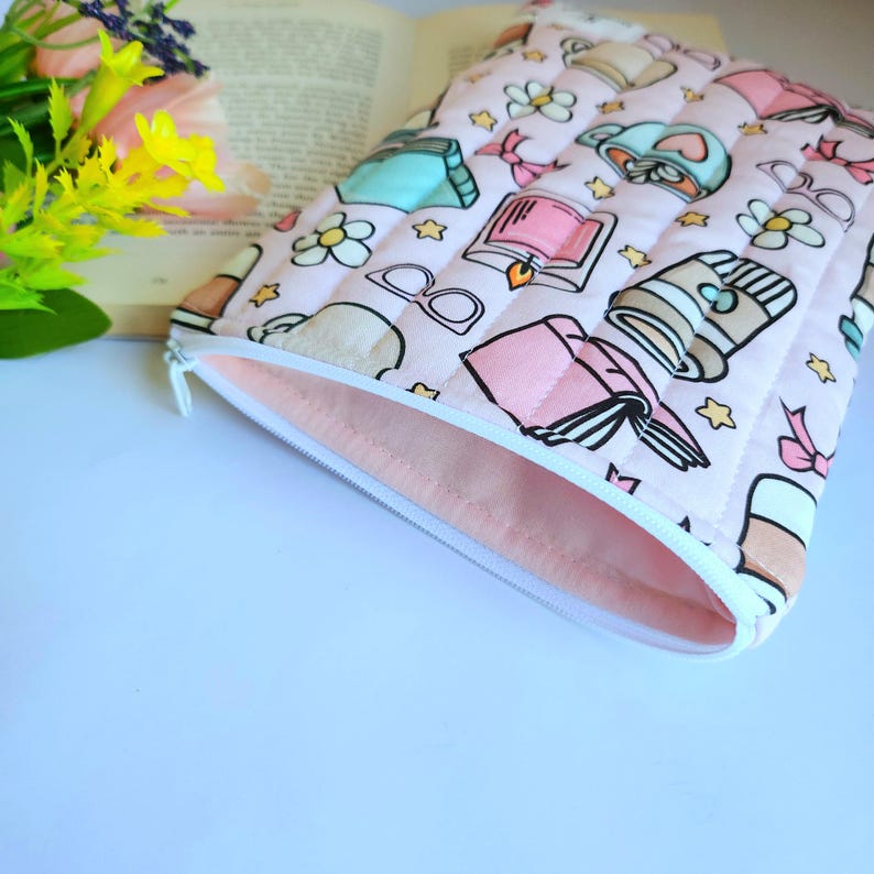 May include: A quilted fabric pouch in a light pink hue featuring a repeating pattern of books, coffee cups, eyeglasses, and small flowers. The rectangular case has a white zipper closure along the top edge and a solid light pink interior lining.