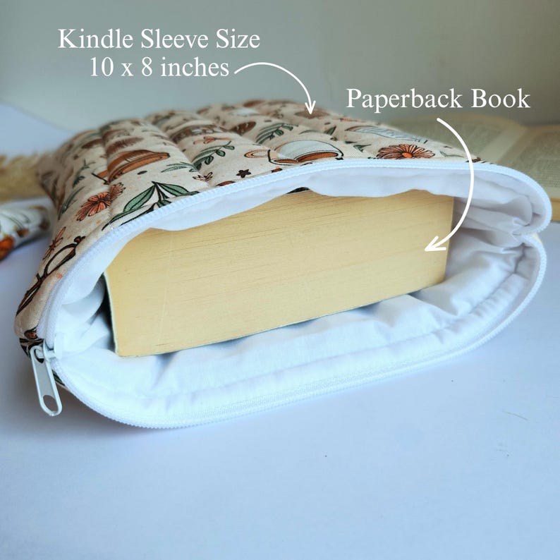 May include: A quilted fabric sleeve with a beige background and a pattern of coffee cups and flowers, containing a paperback book. The sleeve features a white zipper closure and is labeled with the text Kindle Sleeve Size 10 x 8 inches.