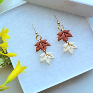 May include: A pair of dangle earrings featuring two stacked maple leaf charms in matte terracotta and cream colors. The leaves are connected by gold-toned metal jump rings and suspended from gold-toned fishhook ear wires for a secure fit.