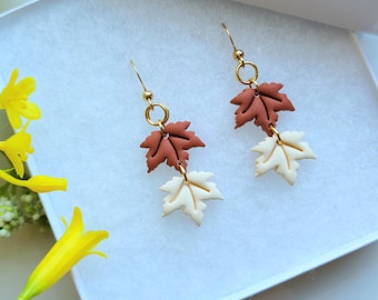 Pode incluir: Um par de brincos pendentes com dois berloques de folha de bordo sobrepostos em cores terracota fosco e creme. As folhas são conectadas por anéis de metal dourado e suspensas por ganchos de orelha em tom dourado.
