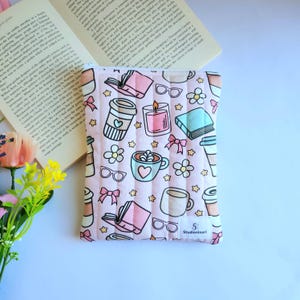 May include: A rectangular quilted fabric pouch featuring a light pink background with a repeating pattern of coffee cups, books, eyeglasses, candles, flowers, and bows. A small white label with the text Studioninori is sewn on the bottom right.