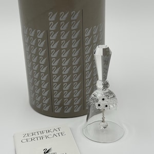 May include: A clear crystal bell with a faceted handle and a small floral accent featuring three white petals and black centers. A small crystal clapper hangs inside. Behind it is a gray cylindrical box labeled SWAROVSKI SILVER CRYSTAL, and a white certificate.
