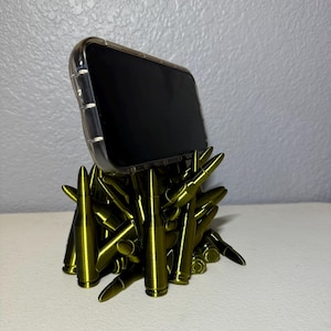 May include: A phone stand made from a cluster of gold-colored bullet casings. The stand is holding a clear phone case with a black phone inside.