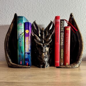 A dragon bookend with a gold finish holding up a variety of books.  'Wings of Fire', Dragon Bookend, Fantasy Bookend, Book Decor, Hand Painted Dragon Bookend, Dragon Shelf Decor, Book Protector Dragon, Fantasy Bookshelf, Antique Gold Dragon Decor, Dragon Bookend with Antique Gold Highlights, Dragon Bookend for Fantasy Lovers, Bookend That Looks Like a Dragon Guarding Books, Decorative Dragon Bookend for Bookshelves, Fantasy Decor Bookend, book lover gifts, book lover, book gift, onyx storm