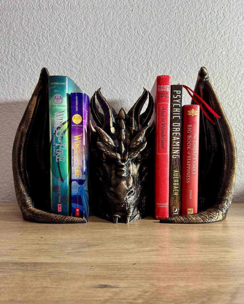 A dragon bookend with a gold finish holding up a variety of books.  'Wings of Fire', Dragon Bookend, Fantasy Bookend, Book Decor, Hand Painted Dragon Bookend, Dragon Shelf Decor, Book Protector Dragon, Fantasy Bookshelf, Antique Gold Dragon Decor, Dragon Bookend with Antique Gold Highlights, Dragon Bookend for Fantasy Lovers, Bookend That Looks Like a Dragon Guarding Books, Decorative Dragon Bookend for Bookshelves, Fantasy Decor Bookend, book lover gifts, book lover, book gift, onyx storm