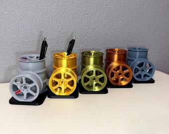 May include: Five different colored desk organizers shaped like car wheels. The organizers are made of plastic and have a black base. The colors are gray, gold, green, copper, and blue.