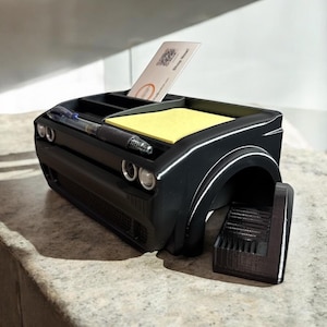 May include: Black desk organizer shaped like the front end of a car. It has a compartment for pens, a sticky note holder, and a card holder. The organizer is made of 3D printed plastic.