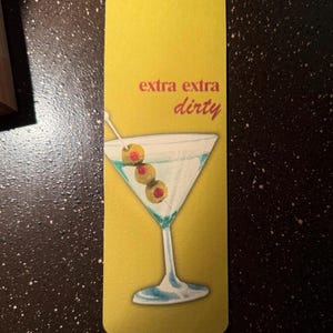 May include: A yellow bookmark with the text "extra extra dirty" in red and a martini glass with olives on a toothpick.