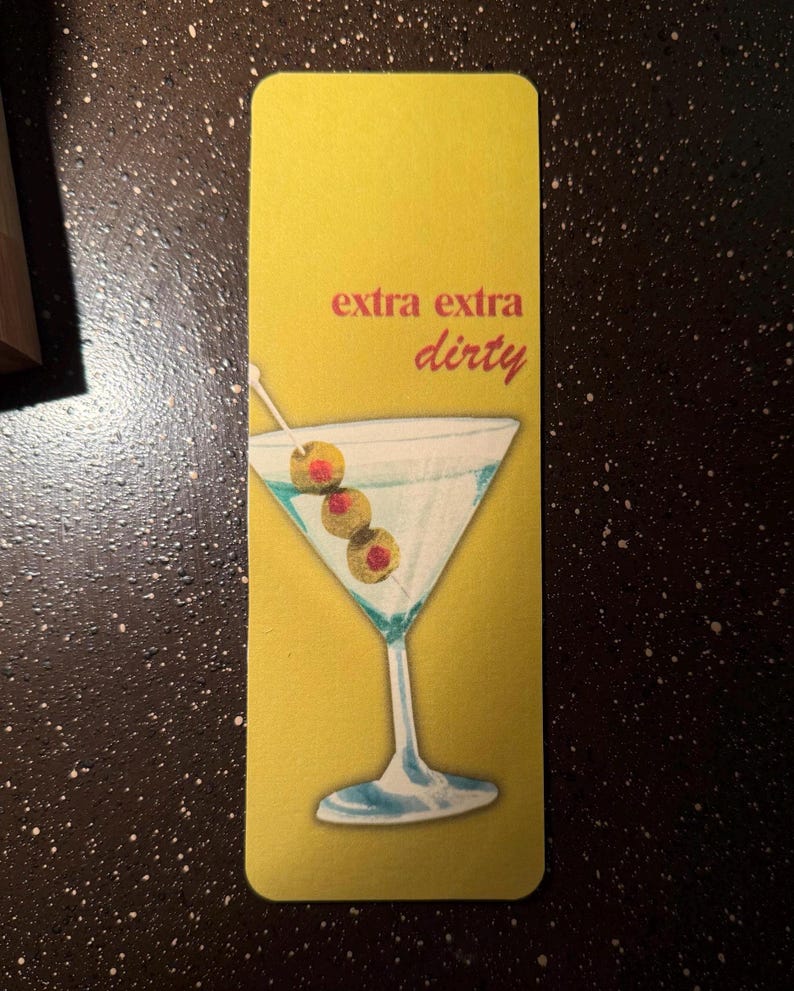 May include: A yellow bookmark with the text "extra extra dirty" in red and a martini glass with olives on a toothpick.
