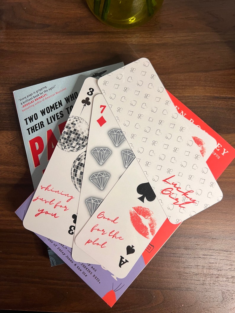 May include: Four bookmarks with a playing card design. The bookmarks feature a red heart, a black spade, a black club, and a diamond. The bookmarks have text that reads "Shining just for you", "Good for the plot", "Lucky Girl", and "7".