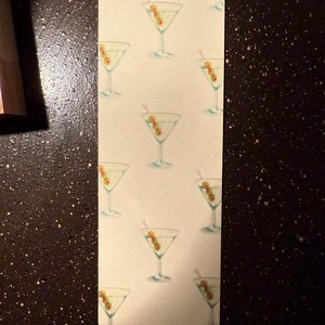 May include: A white bookmark with a repeating pattern of green olives and martini glasses. The glasses are filled with a clear liquid and have a green olive garnish.