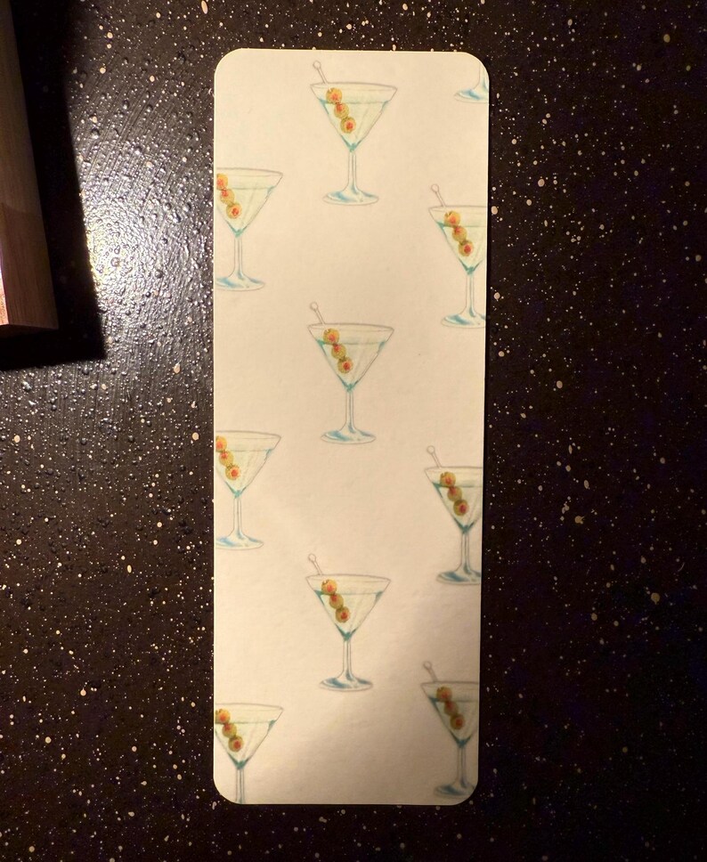May include: A white bookmark with a repeating pattern of green olives and martini glasses. The glasses are filled with a clear liquid and have a green olive garnish.