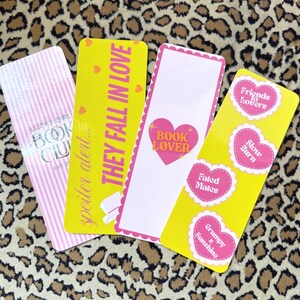 May include: Four yellow bookmarks with pink hearts and text. The bookmarks feature phrases like "They Fall in Love", "Book Lover", "Fated Mates", and "Friends to Lovers".