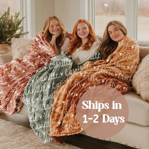 May include: Three personalized blankets in shades of pink, green, and orange are draped over a sofa. Each blanket features a repeating pattern of names in white script. The blankets are soft and cozy, perfect for snuggling.