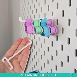 May include: A white pegboard with several multi-colored cookie cutters and a white hook. The cookie cutters are in shades of purple, green, and blue. The hook is held in a hand. The text "3D PRINTING FILES | STL" is at the bottom.