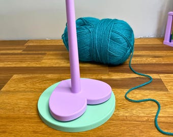 May include: A two-tone yarn holder in light purple and mint green, with a tall central post and a round base. A ball of teal yarn sits behind the holder, with the yarn extending towards the holder. The text "3D PRINTING FILES | STL" is at the bottom.