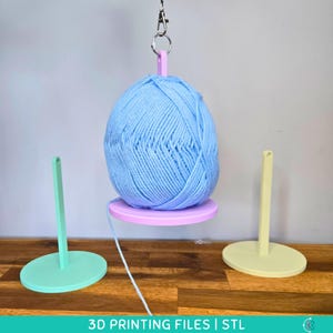 Peut inclure: Une pelote de laine bleu clair suspendue par un support à fil rose imprimé en 3D. Deux autres supports à fil, un vert menthe et un jaune, sont sur une surface en bois. Le texte "3D PRINTING FILES | STL" est en bas.