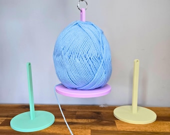 May include: A light blue yarn ball suspended by a pink 3D-printed yarn holder. Two additional yarn holders, one mint green and one yellow, are on a wooden surface. The text "3D PRINTING FILES | STL" is at the bottom.