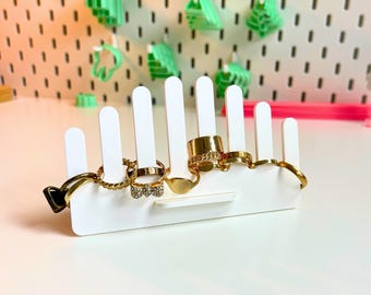 Op de afbeelding: Een witte ringhouder met verticale sleuven met diverse gouden ringen. De ringen omvatten een dikke band, een schakelkettingontwerp en ringen met decoratieve stenen. De achtergrond toont groene boomvormige objecten en een pegboard.