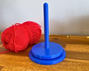May include: A blue yarn swift with a vertical post and a circular base, next to two balls of red yarn. The swift is designed to hold yarn while winding or unwinding. The text "3D PRINTING FILES | STL" is at the bottom.