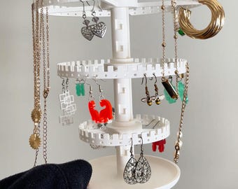 May include: A white, three-tiered jewelry organizer displaying various earrings and necklaces. The organizer has a circular base and rotating shelves with notched edges. The jewelry includes gold hoops, heart-shaped earrings, and long necklaces.