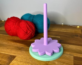 May include: A lilac yarn holder with a flower-shaped base and a mint green bottom. Two balls of red yarn and two balls of teal yarn are behind the holder. The text "3D PRINTING FILES | STL" is at the bottom.