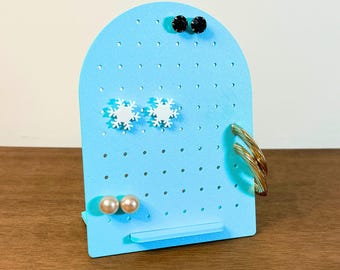 May include: A light blue earring display stand with multiple holes for hanging earrings. The stand holds black stud earrings, white snowflake earrings, and pearl earrings. Gold hoop earrings are also displayed. The text "3D PRINTING FILES | STL" is at the bottom.