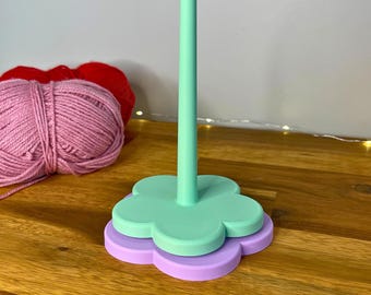 May include: A mint green and purple yarn holder with a flower-shaped base and a central post. Two balls of yarn, one pink and one red, are visible in the background. The text "3D PRINTING FILES | STL" is at the bottom.