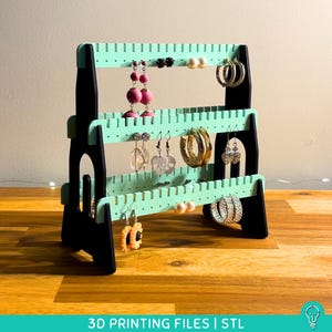 May include: A three-tiered black and mint green earring display stand. The stand holds various earrings, including hoops, studs, and dangling styles. The text "3D PRINTING FILES | STL" is at the bottom of the image.