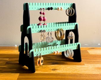 May include: A three-tiered black and mint green earring display stand. The stand holds various earrings, including hoops, studs, and dangling styles. The text "3D PRINTING FILES | STL" is at the bottom of the image.