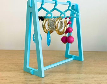 May include: A light blue, 3D-printed jewellery display stand. The stand has miniature hanger-shaped hooks for earrings and other jewellery. Various earrings in gold, silver, and pink are displayed. The text "3D PRINTING FILES | STL" is at the bottom.