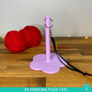 May include: A lilac yarn holder with a flower-shaped base and a silver keyring. A black lanyard is attached to the keyring. Two balls of red yarn are in the background. The text "3D PRINTING FILES | STL" is at the bottom.